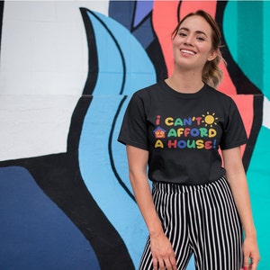 May include: A woman wearing a black t-shirt with the text "I can't afford a house!" in colorful letters and a house and sun graphic. She is standing in front of a colorful wall.
