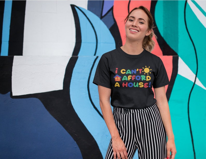 May include: A woman wearing a black t-shirt with the text "I can't afford a house!" in colorful letters and a house and sun graphic. She is standing in front of a colorful wall.