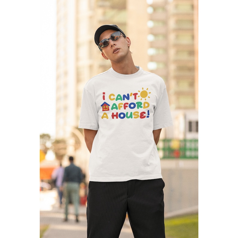 May include: White t-shirt with a colorful graphic that says "I can't afford a house!" The graphic features a house, a sun, and the words in different colors.