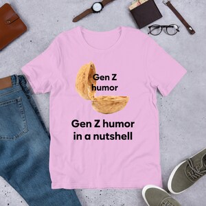 May include: Pink t-shirt with a graphic of a walnut shell with the text "Gen Z humor" written on the top half of the shell and the text "Gen Z humor in a nutshell" written below the shell.