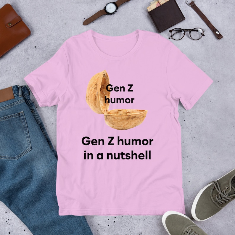 May include: Pink t-shirt with a graphic of a walnut shell with the text "Gen Z humor" written on the top half of the shell and the text "Gen Z humor in a nutshell" written below the shell.
