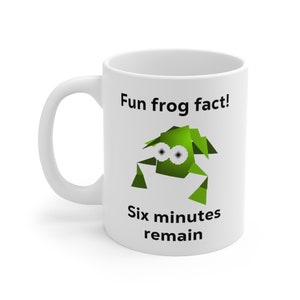 Może przedstawiać: Biały kubek ceramiczny z zieloną kreskówkową żabą i tekstem "Fun frog fact! Six minutes remain".