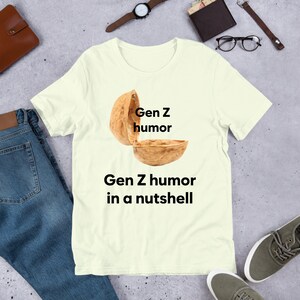 May include: A light yellow t-shirt with a graphic of a walnut shell with the text "Gen Z humor" written on the top half of the shell and the text "Gen Z humor in a nutshell" written below the shell.