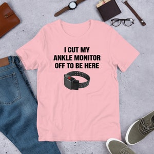 May include: Pink t-shirt with the text "I cut my ankle monitor off to be here" and a black ankle monitor with a red button.