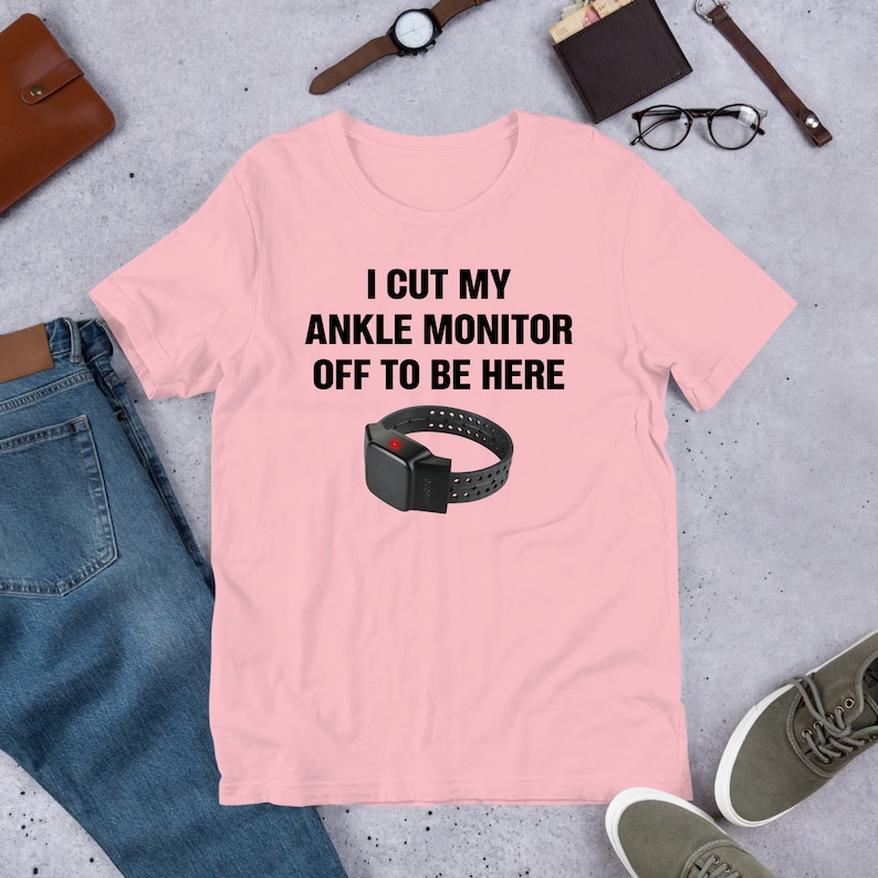 May include: Pink t-shirt with the text "I cut my ankle monitor off to be here" and a black ankle monitor with a red button.