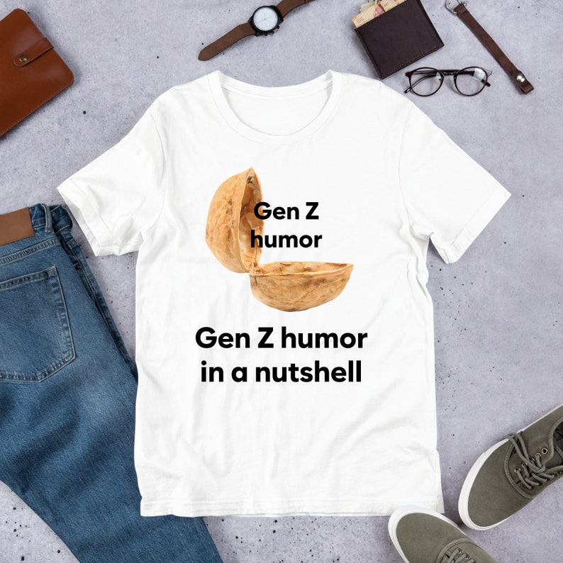 May include: White t-shirt with a graphic of a walnut shell with the text "Gen Z humor" written on the top half of the shell and "Gen Z humor in a nutshell" written below the shell.