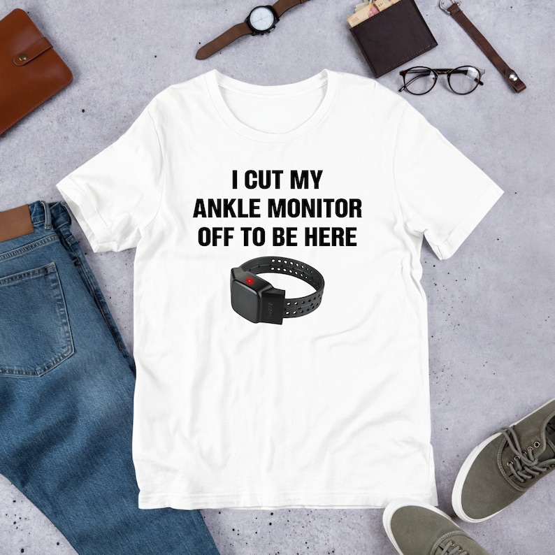 May include: White t-shirt with black text that reads "I cut my ankle monitor off to be here". A black ankle monitor with a red light is pictured below the text.