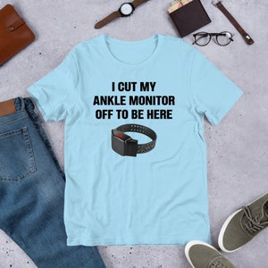 May include: A light blue t-shirt with the text "I cut my ankle monitor off to be here" in black. A black ankle monitor with a red button is pictured below the text.