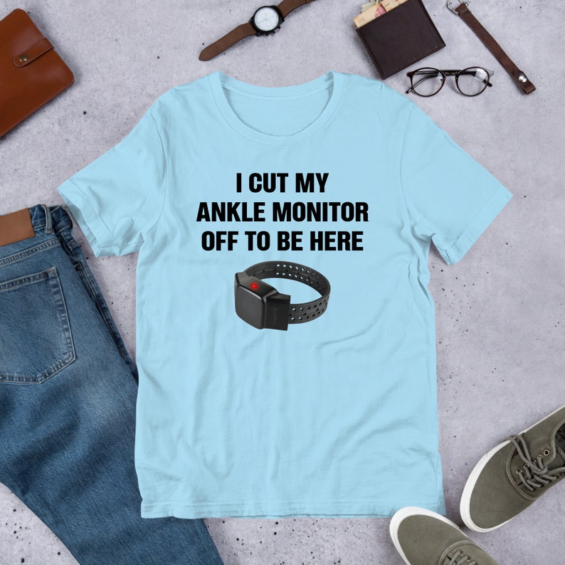 May include: A light blue t-shirt with the text "I cut my ankle monitor off to be here" in black. A black ankle monitor with a red button is pictured below the text.