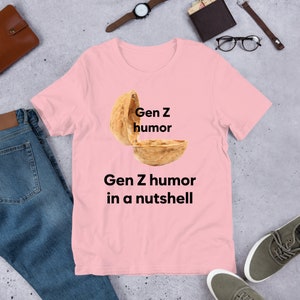 May include: Pink t-shirt with a graphic of a walnut shell with the text "Gen Z humor" written on the top half of the shell and the text "Gen Z humor in a nutshell" written below the shell.