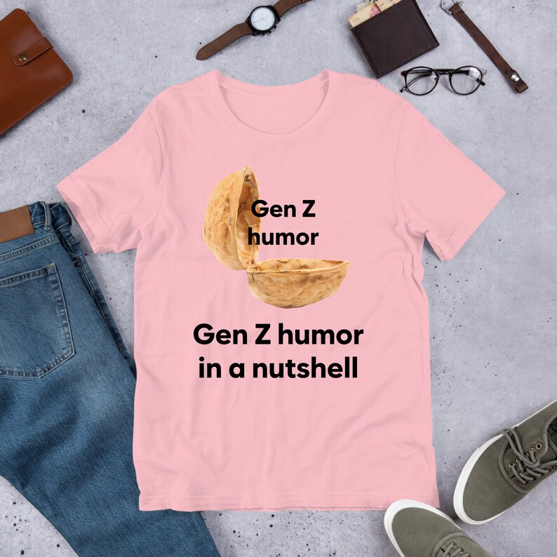 May include: Pink t-shirt with a graphic of a walnut shell with the text "Gen Z humor" written on the top half of the shell and the text "Gen Z humor in a nutshell" written below the shell.