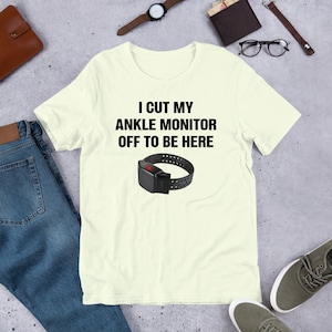 May include: A light yellow t-shirt with the text "I cut my ankle monitor off to be here" and a black ankle monitor with a red light on it.