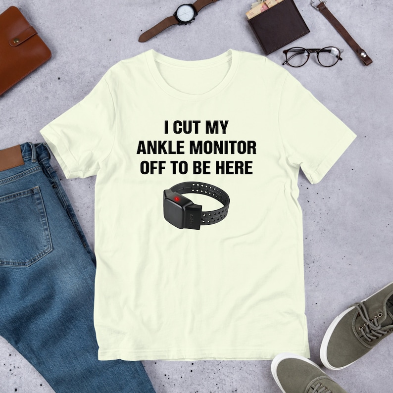 May include: A light yellow t-shirt with the text "I cut my ankle monitor off to be here" and a black ankle monitor with a red light on it.