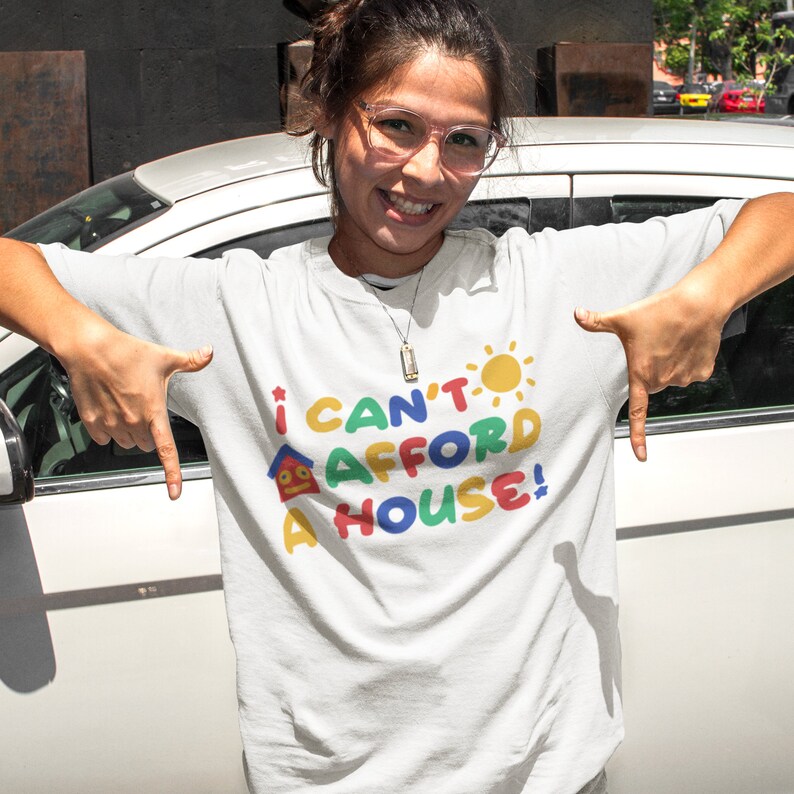 May include: White t-shirt with a colorful graphic that says "I can't afford a house!" The graphic features a house, a sun, and a star.