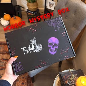 以下が含まれることがあります： 赤い滴るテキストで「HORROR MYSTERY BOX」という言葉が入った黒い箱。箱には紫色のドクロ、クモの巣、「Twisted Retreat」のロゴが描かれています。箱は人の手で持たれています。