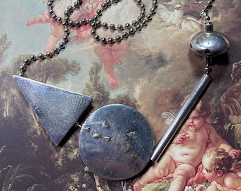 May include: Silver-toned necklace featuring geometric shapes: a triangle, a circle with small triangle accents, and a long, thin bar. The necklace has a ball chain and a decorative, rounded pendant. The background is a classical painting.