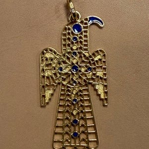May include: Gold-toned pendant in the shape of a bird with intricate openwork design. The pendant features blue enamel accents and circular blue gemstones. The bird has a curved beak and outstretched wings, creating a symmetrical design.