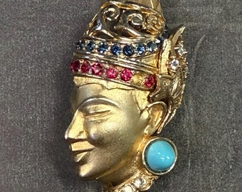 May include: A gold-toned brooch featuring a stylized face with a turquoise stone earring and rhinestones. The face is adorned with red and blue gemstones.