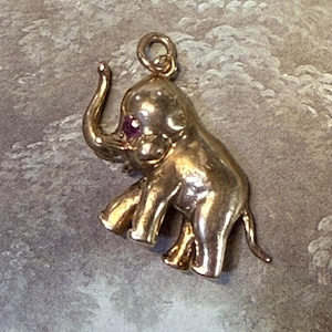 May include: A gold-colored elephant charm with a pink gemstone eye. The charm is detailed with a raised trunk and legs.