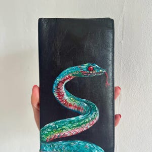 May include: Black leather wallet with a hand-painted snake design. The snake is teal, green, and red, with a red tongue and eyes. The rectangular wallet is held against a white background. The wallet is approximately 20 cm in length.