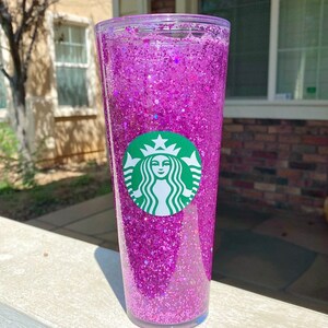 May include: A clear tumbler with a pink glitter design and a green Starbucks logo.
