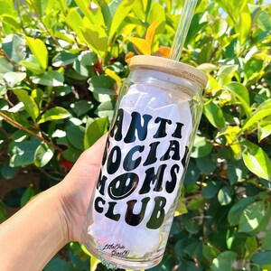 May include: A clear glass water bottle with a bamboo lid and a black vinyl decal that says "Anti Social Moms Club" with a smiley face. The bottle has a glass straw.