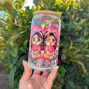 May include: Clear glass can-shaped tumbler with a bamboo lid. The tumbler features a pink heart design with two cartoon girls, each with a pink shirt and crying. The text "Little Chic Shop Co" is on the lid.