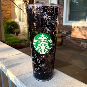 May include: A clear plastic tumbler with a black glitter design and a green Starbucks logo.