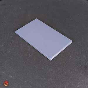 Puede incluir: Una superficie rectangular texturizada de color púrpura claro. La superficie tiene un borde ligeramente elevado y un patrón moteado. El objeto está sobre un fondo gris oscuro.