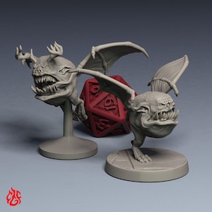 May include: Two grey unpainted miniature figures of winged creatures with large mouths, sharp teeth, and antler-like protrusions on their heads. Each figure stands on a circular base, positioned in front of a large red twenty-sided gaming die.