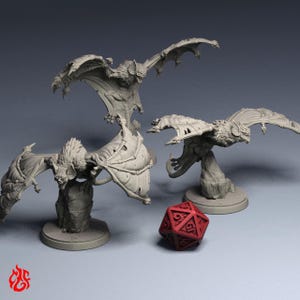 May include: Three unpainted grey miniature bat creatures with outstretched wings, each mounted on a circular base, positioned alongside a single red twenty-sided die. The figures feature detailed fur textures and skeletal wing structures.
