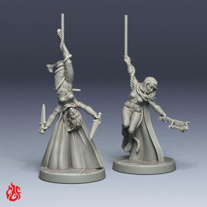 May include: Two unpainted grey resin miniature figurines. One figure hangs upside down from a rope, holding two daggers. The other figure is suspended upright by a rope, wearing a hooded cloak and holding a crossbow. Both stand on round bases.