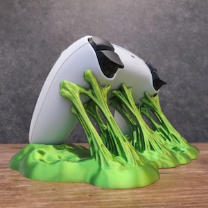 Pode incluir: Um suporte verde impresso em 3D para um controlador de videogame. O suporte tem a forma de uma série de videiras ou galhos. O controlador é branco com botões pretos.
