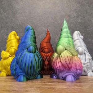Könnte beinhalten: Fünf Gnome, jeder in einer anderen Farbe bemalt, stehen in einer Reihe auf einer Holzoberfläche. Die Gnome sind gelb, blau und grün, rot und silber.