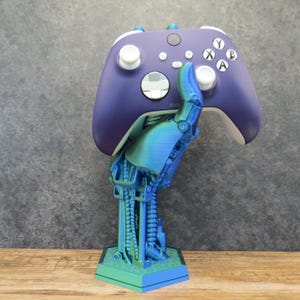 Könnte beinhalten: Eine 3D-gedruckte Roboterhand hält einen lila und weißen Xbox-Controller. Die Roboterhand ist blau und grün und hat eine sechseckige Basis.