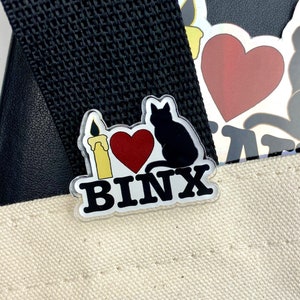 May include: A clear plastic pin with a black cat, a red heart, a yellow candle, and the text "I love Binx".