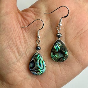 Könnte beinhalten: Ein Paar tropfenförmige Ohrringe mit schillernden Abalone-Muscheln in Grün, Blau und Schwarz. Die Ohrringe haben silberfarbene Haken und eine kleine, dunkelblaue Perle. Die Muscheln haben ein Wirbelmuster.