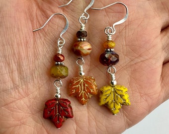 Puede incluir: Tres pendientes colgantes con ganchos plateados y detalles de alambre. Cada pendiente muestra una cuenta facetada y un dije en forma de hoja en colores otoñales: uno rojo, uno naranja con vetas marrones y uno amarillo brillante con detalles dorados.