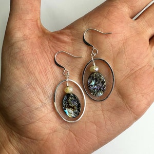 May include: A pair of silver hoop earrings with a round abalone shell pendant and a small white bead. The earrings are hanging from a person's hand.