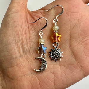 Pode incluir: Um par de brincos de prata com um charme de lua crescente, um charme de estrela e um charme de sol. Os brincos são acentuados com contas de vidro iridescente e âmbar.