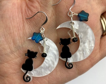 May include: A pair of silver earrings featuring a white crescent moon with a black cat silhouette sitting on it. Each earring has a blue star charm and a clear crystal star charm.