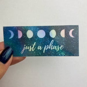 May include: A sticker with a galaxy background and a row of iridescent moons. The text "just a phase" is written in a white, cursive font.