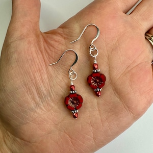 May include: A pair of red flower earrings with silver hooks. Each earring has a red flower-shaped bead, with smaller red and silver beads. The earrings are shown against a light background.