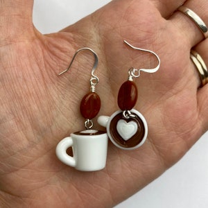 May include: Coffee-themed earrings featuring white ceramic coffee cups with brown coffee and a white heart design. Each earring has a brown coffee bean bead and silver-colored ear hooks. A charming accessory for coffee lovers.