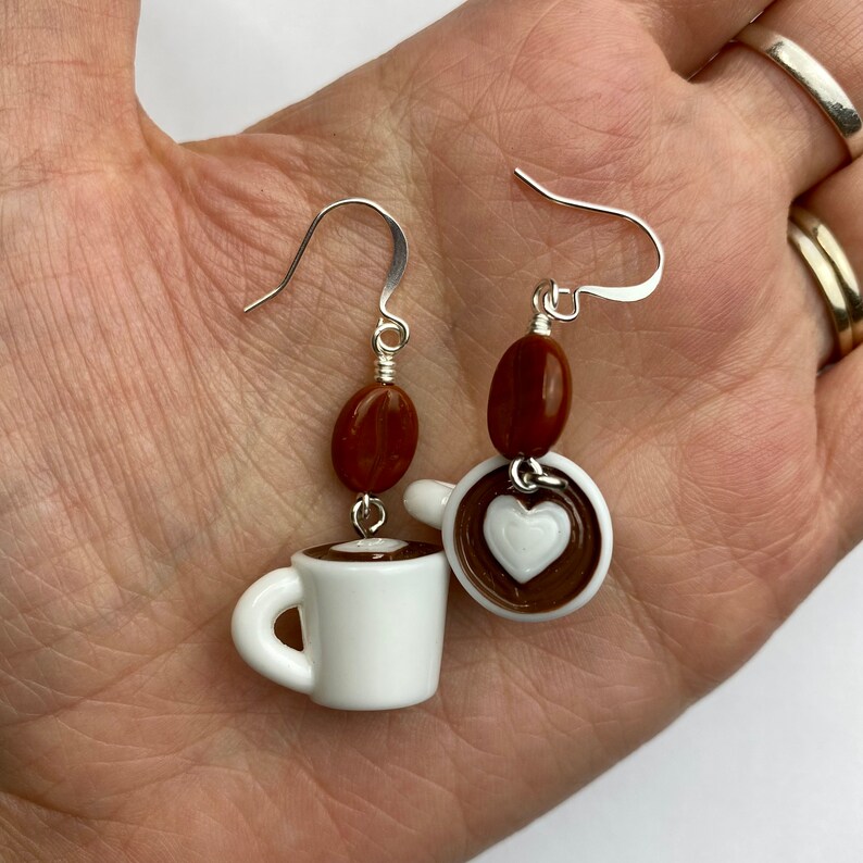 May include: Coffee-themed earrings featuring white ceramic coffee cups with brown coffee and a white heart design. Each earring has a brown coffee bean bead and silver-colored ear hooks. A charming accessory for coffee lovers.