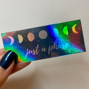 May include: A holographic sticker with a black background and a rainbow glitter effect. The sticker features a series of moons in different phases and the text "just a phase".