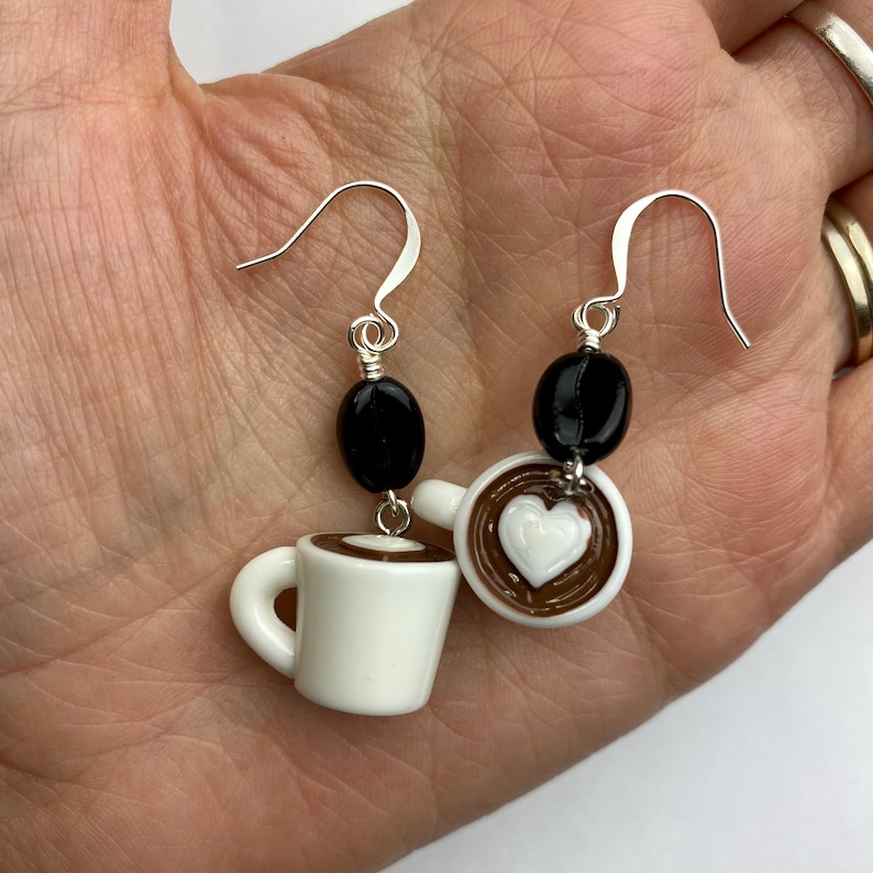 May include: A pair of coffee-themed earrings. Each earring features a white ceramic coffee cup with a brown rim and a black coffee bean. One cup has a heart-shaped design in the coffee. The earrings have silver-colored ear wires.