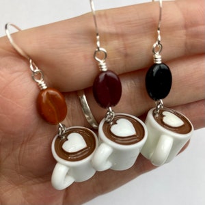May include: Silver dangle earrings featuring coffee-themed charms. Each earring has a white coffee cup charm with a heart design, paired with a coffee bean or oval bead in shades of brown and black. A charming accessory for coffee lovers.