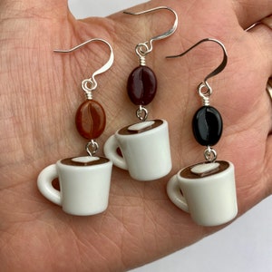 May include: Three pairs of coffee-themed earrings. Each earring features a small white coffee cup charm with a brown coffee design, paired with a coffee bean bead. The earrings have silver-colored hooks and wire detailing.