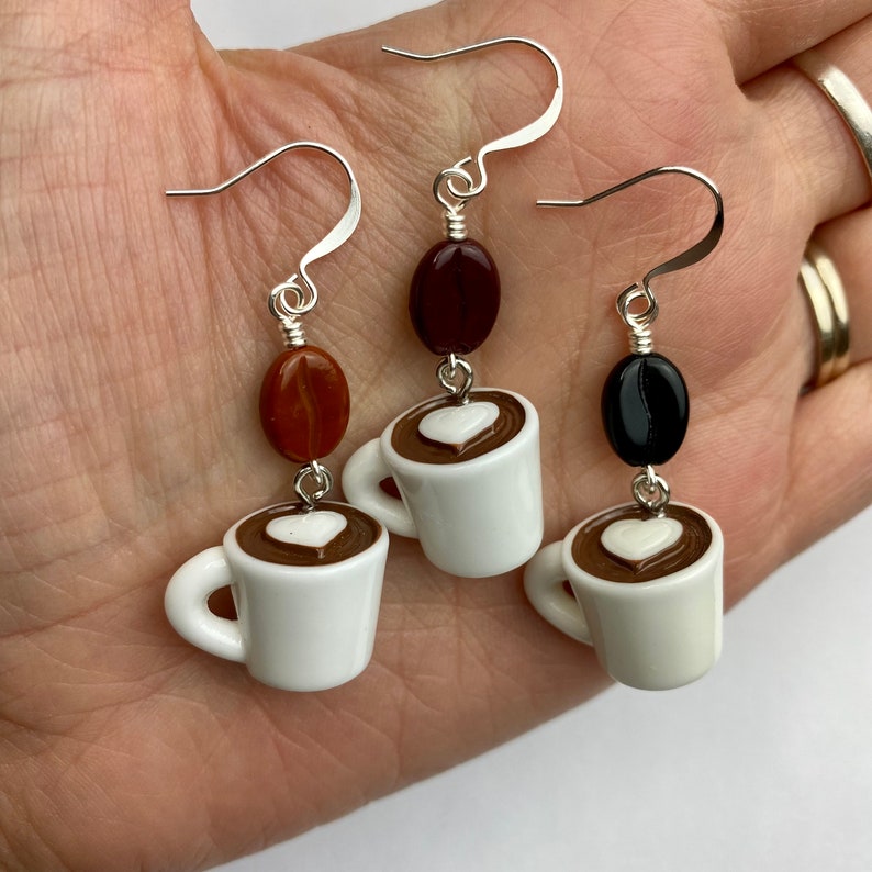 May include: Silver-tone dangle earrings featuring coffee-themed charms. Each earring has a silver hook, a coffee bean bead, and a white mug charm with a heart-shaped design in the coffee. The earrings are a unique accessory for coffee lovers.